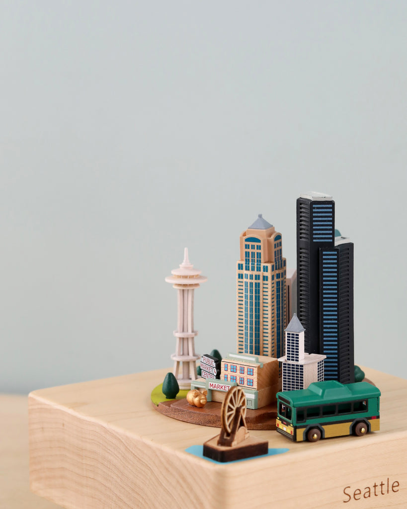 The Seattle Wooden Music Box showcases a miniaturized diorama of Seattle on a wooden platform, featuring the iconic Space Needle, towering skyscrapers, the Pike Place Market sign, and a green trolley bus. A Ferris wheel is positioned near the front. The wooden base has "Seattle" engraved on it, making it an ideal gift or keepsake.