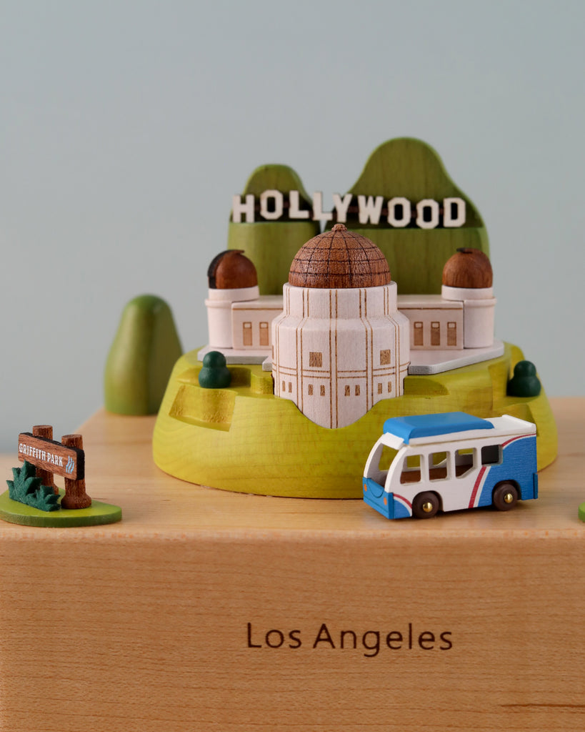 The Los Angeles Wooden Music Box features an intricately designed mini diorama of iconic Los Angeles landmarks, including the Griffith Observatory, the Hollywood sign on a hill, a Griffith Park sign, and a colorful bus. Perfect as a keepsake or gift, this unique piece has "Los Angeles" engraved on its base.