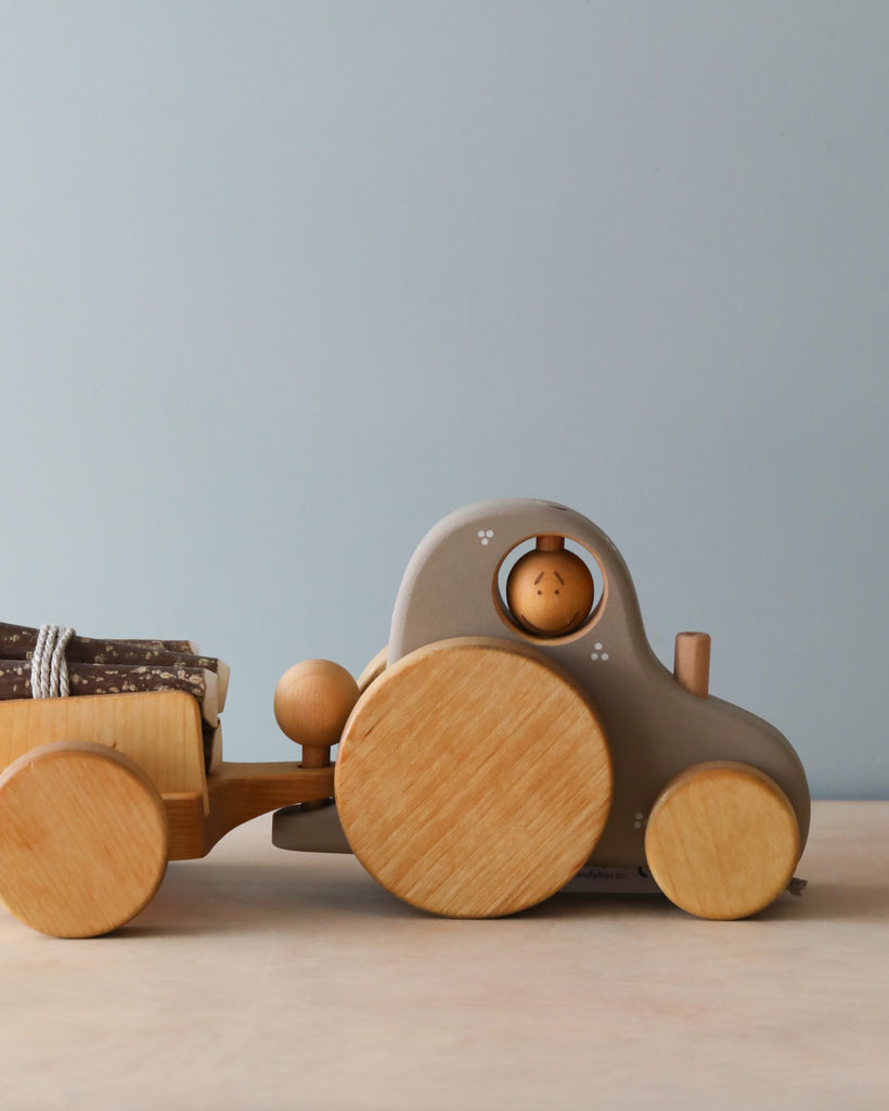 A **Handmade Wooden Tractor** with large circular wheels and a small driver figure inside pulls a log trailer with miniature logs secured by a rope. The detachable trailer adds versatility to this charming toy, displayed against a simple, light-colored background.