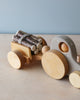 A Handmade Wooden Tractor with smooth, rounded edges and natural tones carries small cut logs tied together in its detachable trailer. The tractor features a smiling wooden figure in the driver's seat and is placed against a light-colored surface with a neutral backdrop.