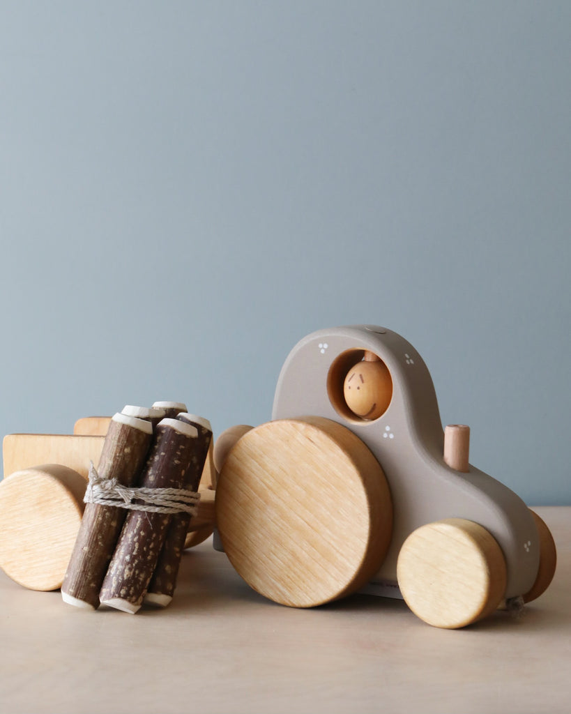 A Handmade Wooden Tractor with a rounded design is shown hauling a detachable trailer filled with small logs tied together. The tractor is a light beige color with wooden wheels, and a small wooden figure sits in the driver's seat. The background is a plain light gray.