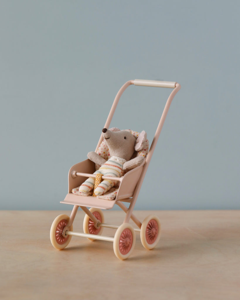 A small stuffed mouse toy dressed in striped clothing sits in a tiny pastel pink Maileg Miniature Stroller, Micro - Powder. The stroller is placed on a light wooden surface against a plain light blue background.