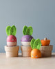 Three colorful, round toys resembling vegetables with leaves on top, each sitting on a small wooden cup. They have smiling faces—one pink, one purple, and one orange. Perfect for budding gardeners, these educational wooden toys include another piece resembling a halved orange vegetable beside them. Stacking Wooden Veggies