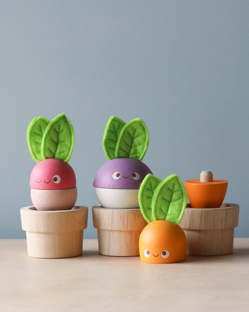 Three colorful, round toys resembling vegetables with leaves on top, each sitting on a small wooden cup. They have smiling faces—one pink, one purple, and one orange. Perfect for budding gardeners, these educational wooden toys include another piece resembling a halved orange vegetable beside them. Stacking Wooden Veggies