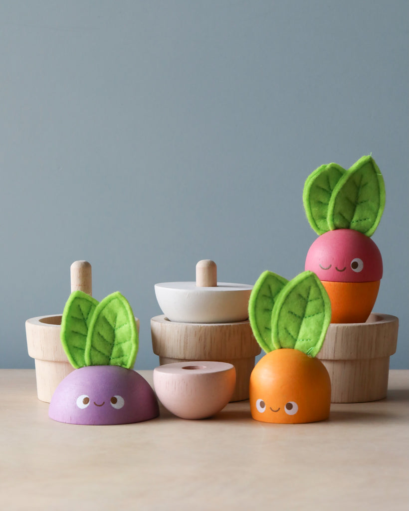 Stacking Wooden Veggies with smiling faces and green felt leaves are displayed on a table, perfect for budding gardeners. The toys come in various shapes and colors like purple, orange, and pink. Wooden bowls with lids are seen in the background against a light blue wall, adding an educational toy element to playtime.