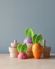 A group of Stacking Wooden Veggies, perfect for budding gardeners, includes a pink radish with green felt leaves, a purple turnip, and an orange carrot with smiley faces. They are set against a neutral background inside a wooden basket on a wooden surface.