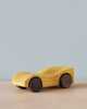 A smooth, yellow Handmade Wooden Race Car - Yellow made from alder with four black wheels rests on a light wooden surface against a plain, light blue background. Coated in non-toxic paint, the race car has a minimalist design without details like windows or lights.