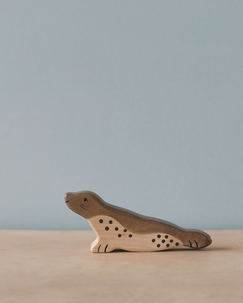 A handcrafted wood toy resembling a seal, with a light brown body, darker brown spots, and simple facial features sits on a light wooden surface against a light blue background. Made in Europe by Holztiger Seal, Head Forward figures, it embodies the charm of quality artistry.