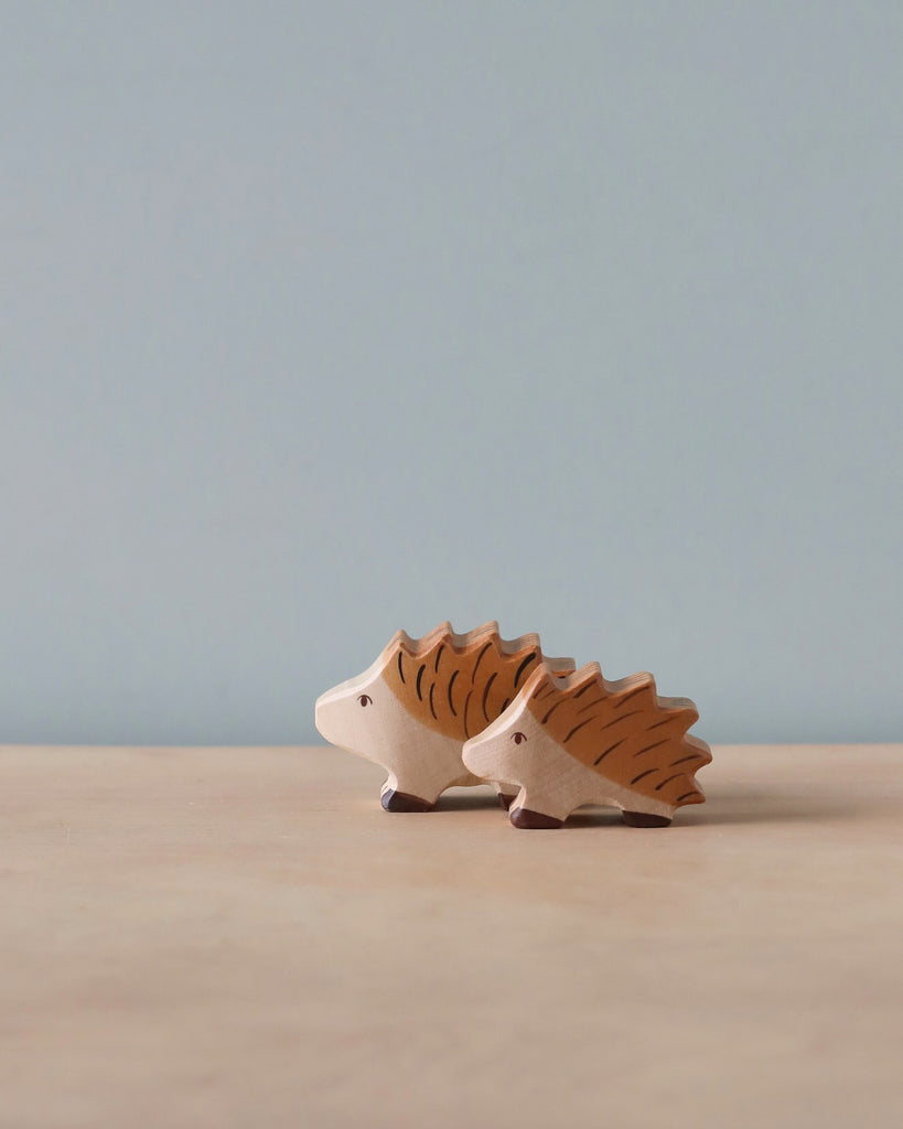 Two Holztiger Hedgehog hedgehog figurines, one smaller than the other, are placed on a light wooden surface with a light blue background. The handcrafted wood figures are light-colored with brown detailing on their backs, resembling quills.