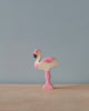 A high-quality Holztiger Flamingo, handcrafted in pink and white, stands gracefully on a flat, light-colored surface against a plain grayish-blue background. This beautiful piece is part of the renowned HOLZTIGER figures collection.