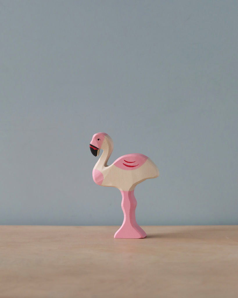 A high-quality Holztiger Flamingo, handcrafted in pink and white, stands gracefully on a flat, light-colored surface against a plain grayish-blue background. This beautiful piece is part of the renowned HOLZTIGER figures collection.