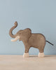 A high-quality Holztiger Elephant, Trunk Raised stands on a light-colored surface against a light blue background. Handcrafted from wood, typical of HOLZTIGER figures, the elephant is painted gray with light-colored tusks, feet, and a black tail. The figure has minimal details and a smooth finish.