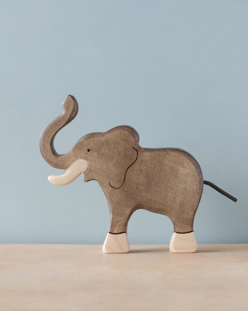 A high-quality Holztiger Elephant, Trunk Raised stands on a light-colored surface against a light blue background. Handcrafted from wood, typical of HOLZTIGER figures, the elephant is painted gray with light-colored tusks, feet, and a black tail. The figure has minimal details and a smooth finish.