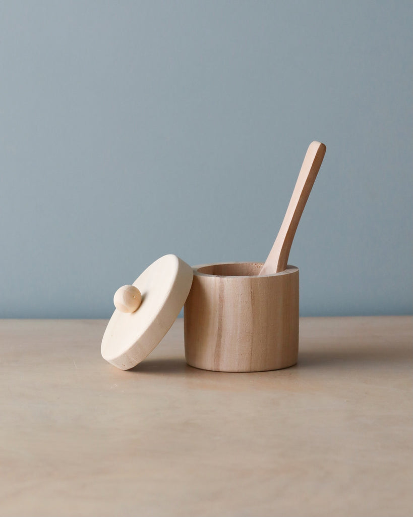 A small Wooden Doll feeding set with a lid and a matching wooden spoon, set on a table against a pale blue background. The lid, adorned with a round knob, rests partially off the pot.