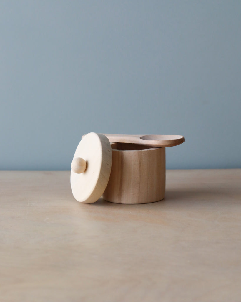 A small handmade Wooden Doll Feeding Set constructed from natural materials, with a circular lid featuring a simple knob on top, positioned on a plain surface against a pale blue background.