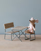 A Maileg Garden Table Set sitting on a miniature chair next to a small table and bench, all crafted from FSC certified wood, set against a plain blue wall on a wooden surface.