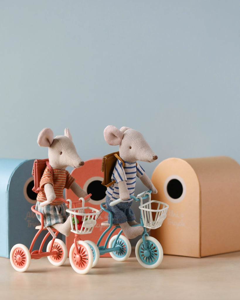 Two fabric mice with magnetic hands ride Maileg Tricycles With Baskets in front of pastel-colored toy houses. The mice are dressed in casual clothes - one in a striped shirt and jeans, the other in a red checked shirt and shorts. The scene is set against a light blue background, complete with wooden seats on their tricycles.