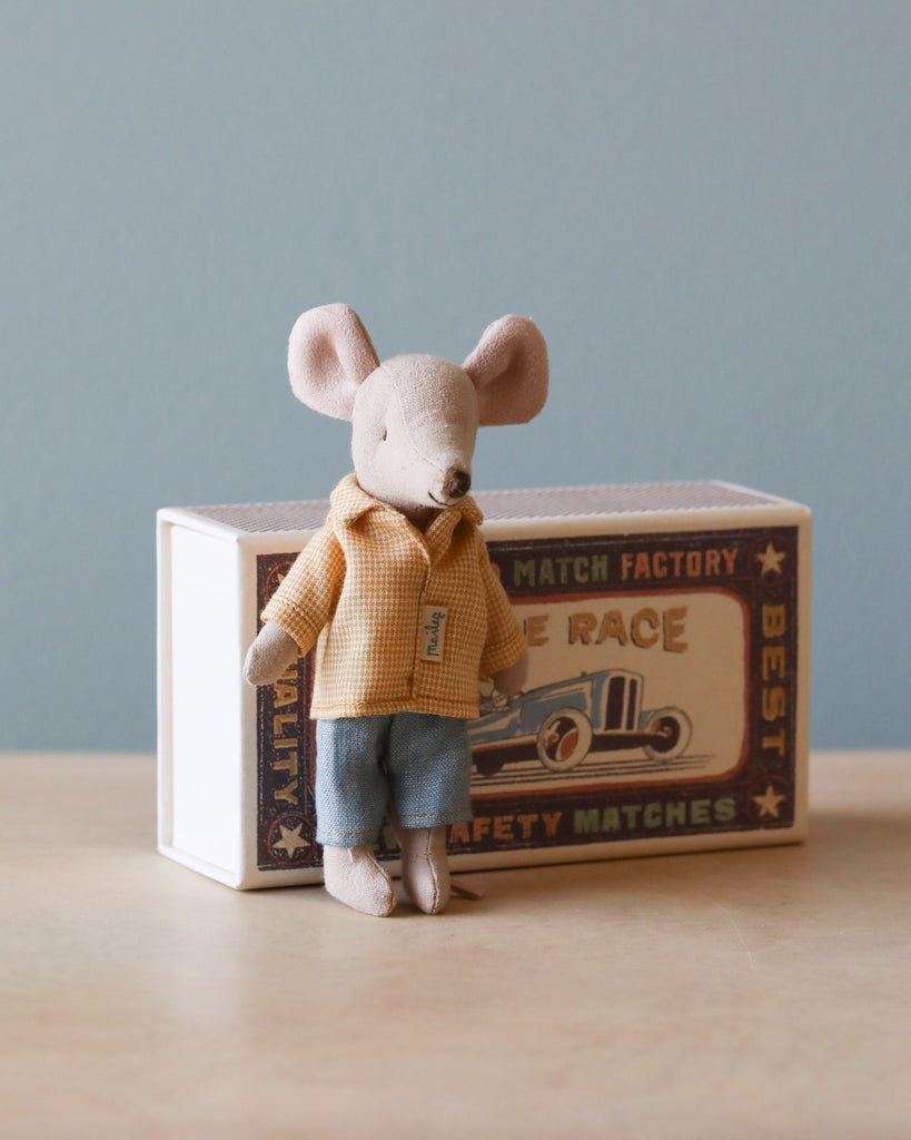 A Maileg Mouse in a box - Big Brother dressed in a cotton yellow checkered shirt and blue shorts, standing beside a vintage matchbox with the text "the race" on a wooden table.