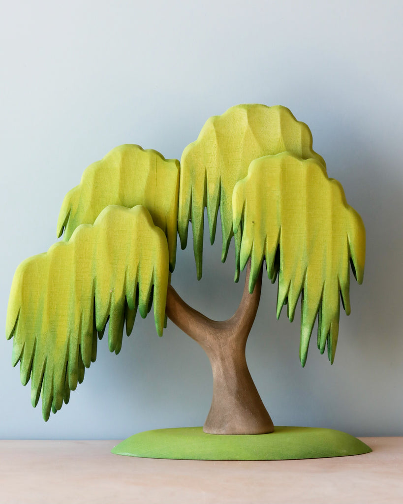 A handmade wooden toy, this Large Wooden Willow Tree stands on a smooth, light green base. The tree has a simple, curved trunk and drooping branches with layered, leaf-like shapes painted in shades of yellow and light green using non-toxic paint, against a plain light gray background.