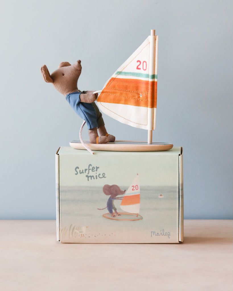 A small toy mouse dressed in blue overalls and a wetsuit stands on a toy sailboard with an orange and green sail marked with the number "20." The mouse, equipped with magnetic hands, is placed on a box labeled "Maileg Little Brother Surfer Mouse," which features an illustration of a mouse on a sailboard.