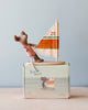 A Maileg Little Sister Surfer Mouse wearing a pink outfit stands on a sailboat with a white sail featuring red, green, and orange stripes with the number 20. The sailboat is placed on a rectangular box that has an illustration of a mouse on a similar sailboat and the text "Surfer Mice." This charming surfing toy even includes magnetic hands for added fun.