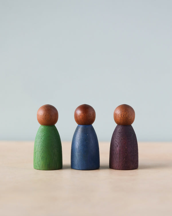 Three Grapat Dark Cold Nins crafted from sustainably forested wood, painted in green, blue, and purple with round brown tops, standing in a row against a soft blue background.