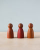 Three Grapat Dark Warm Nins standing in a row against a soft blue background. The center figurine is painted red, while the other two retain a natural wood finish from sustainably forested Sapeli.