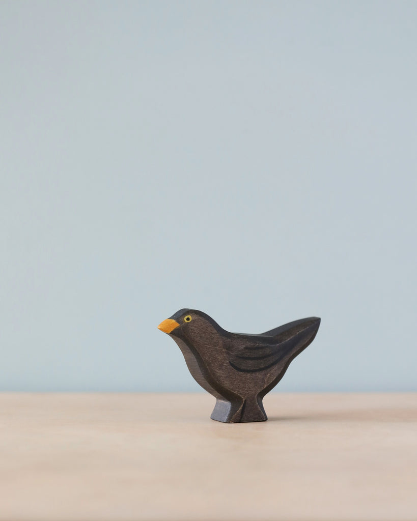 A Handmade Holzwald Blackbird, crafted from sustainable wood, with a black body and a yellow beak, standing on a flat surface against a plain light blue background.