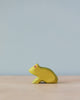 A simple Handmade Holzwald Frog toy painted in bright yellow with natural dyes and a red eye, placed against a soft blue background on a light surface. The frog is in a crouched position, facing.