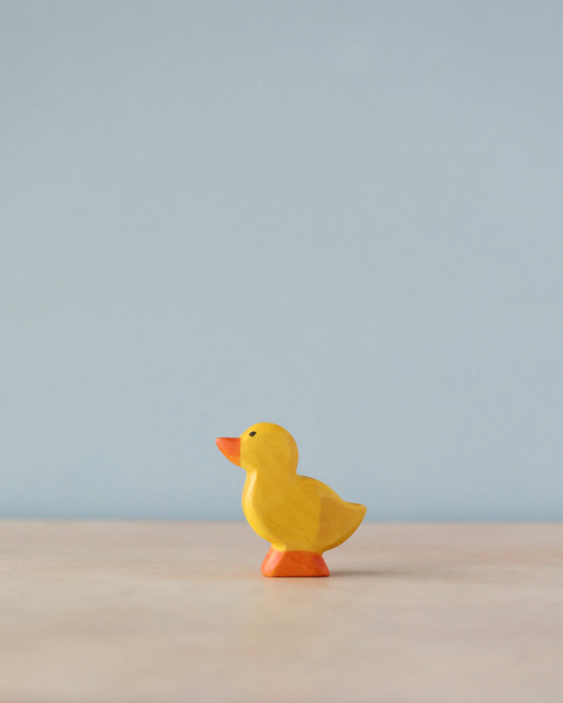 A small, yellow, high-quality Handmade Holzwald Duckling figurine stands on a solid surface against a soft blue background. The duck is profiled to the left with a simple, cartoonish design.