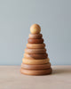 A wooden stacking toy with wooden rings in various natural shade of wood, stacked from large to small, topped with a sphere shaped wooden piece.