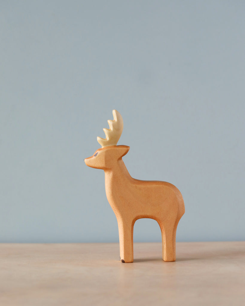 A small wooden Handmade Holzwald Male Deer figurine with detailed antlers stands against a soft blue background. The figurine is simplistic in style, showcasing smooth lines and a light, natural.