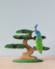 A Handmade Wooden Peacock On A Tree perched on a stylized handmade bonsai tree, with green and blue hues dominating the design. Crafted from lime wood and painted with non-toxic paints, the peacock's tail features multiple eye-like patterns. The background is a plain light blue wall, resting on a light wood surface.