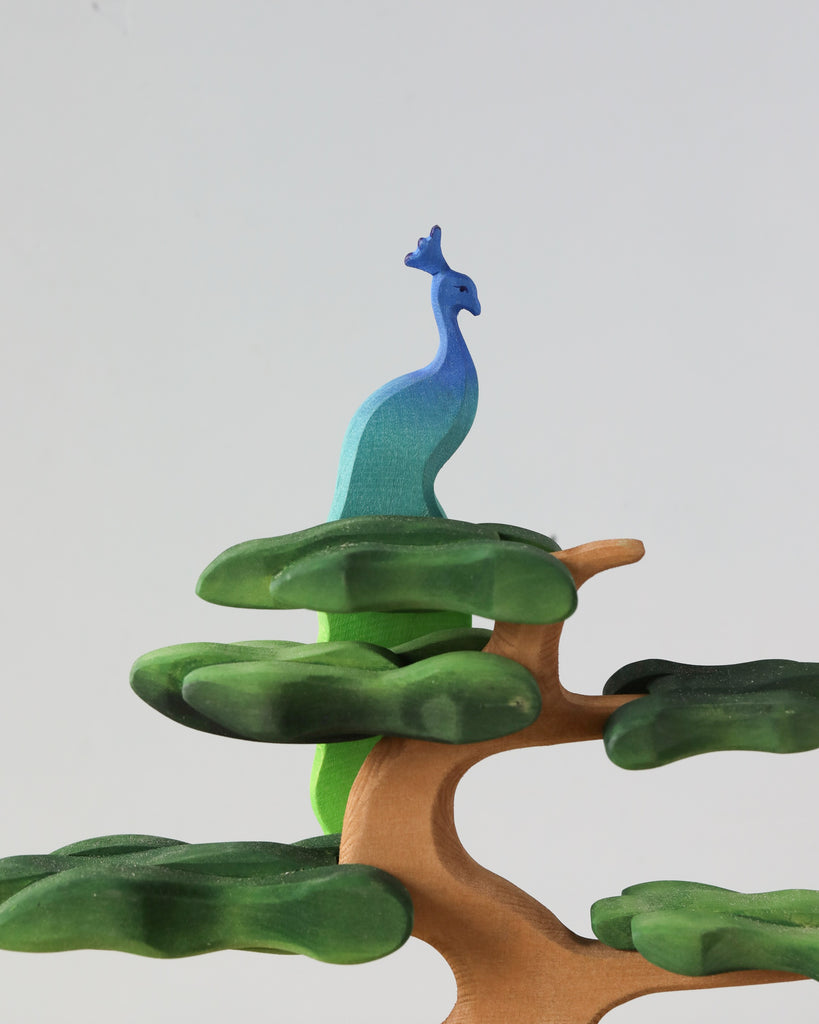 A Handmade Wooden Peacock On A Tree featuring a wooden peacock painted in shades of blue and green is perched atop a stylized tree with broad, green, abstract foliage. Crafted from lime wood and using non-toxic paints, the design is set against a plain light background to highlight its vibrant colors.