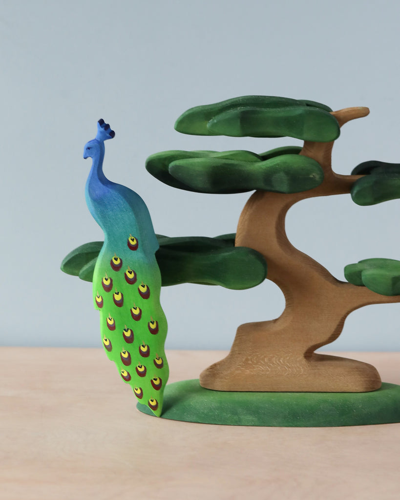 A Handmade Wooden Peacock On A tree with a gradient blue to green body and feather details stands on a small base beside a stylized green wooden tree, crafted from lime wood, with a curvy trunk and flat, rounded leaves. The background is a plain light blue wall.