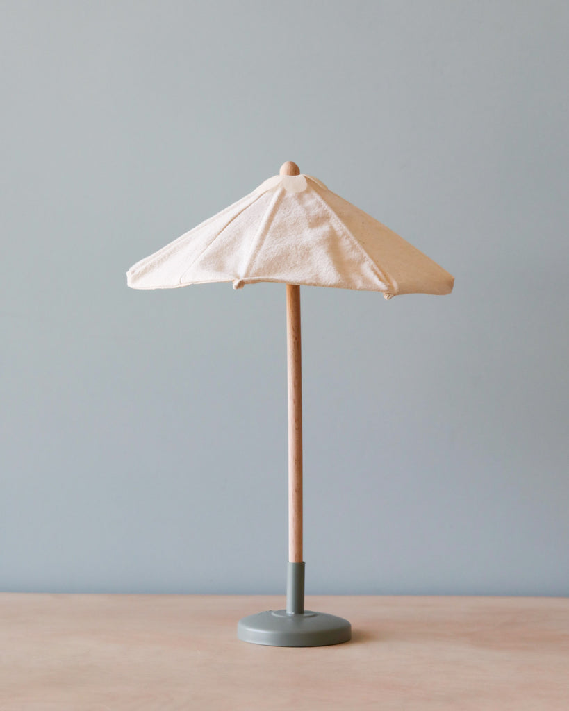 A small beige fabric Maileg Miniature Sunshade with a wooden pole and a gray base stands on a light wood surface, evoking a cozy atmosphere against a light gray background. Perfect for complementing your terrace or miniature garden set, the parasol is folded and has a simple, minimalist design.