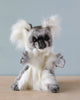 A fluffy Koala Puppet with white and grey fur stands upright, showcasing its large, fuzzy ears. With realistic features, it appears to be waving against a plain, light blue wall that adds a soft contrast to the koala's fur. This delightful puppet is perfect for any cuddly collection.