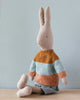 A Maileg Rabbit size 5, Classic - Sweater and Shorts made from recycled polyester is sitting upright against a light blue background. The rabbit is wearing a striped sweater in shades of brown, teal, and orange, along with a gray skirt. Positioned to the side, it displays its long ears and simple facial features.