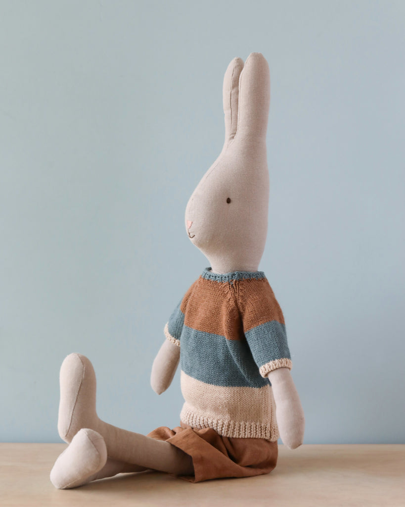 The Maileg Rabbit Size 4, Classic - Knitted Shirt & Shorts is seated against a light blue background. Part of the "bunny and rabbit collection," it wears a blue, beige, and brown striped sweater paired with brown shorts. Made from natural linen and cotton fabric, the rabbit features long ears and a simple, stitched face with a small nose and eyes. Its legs are stretched out in front.