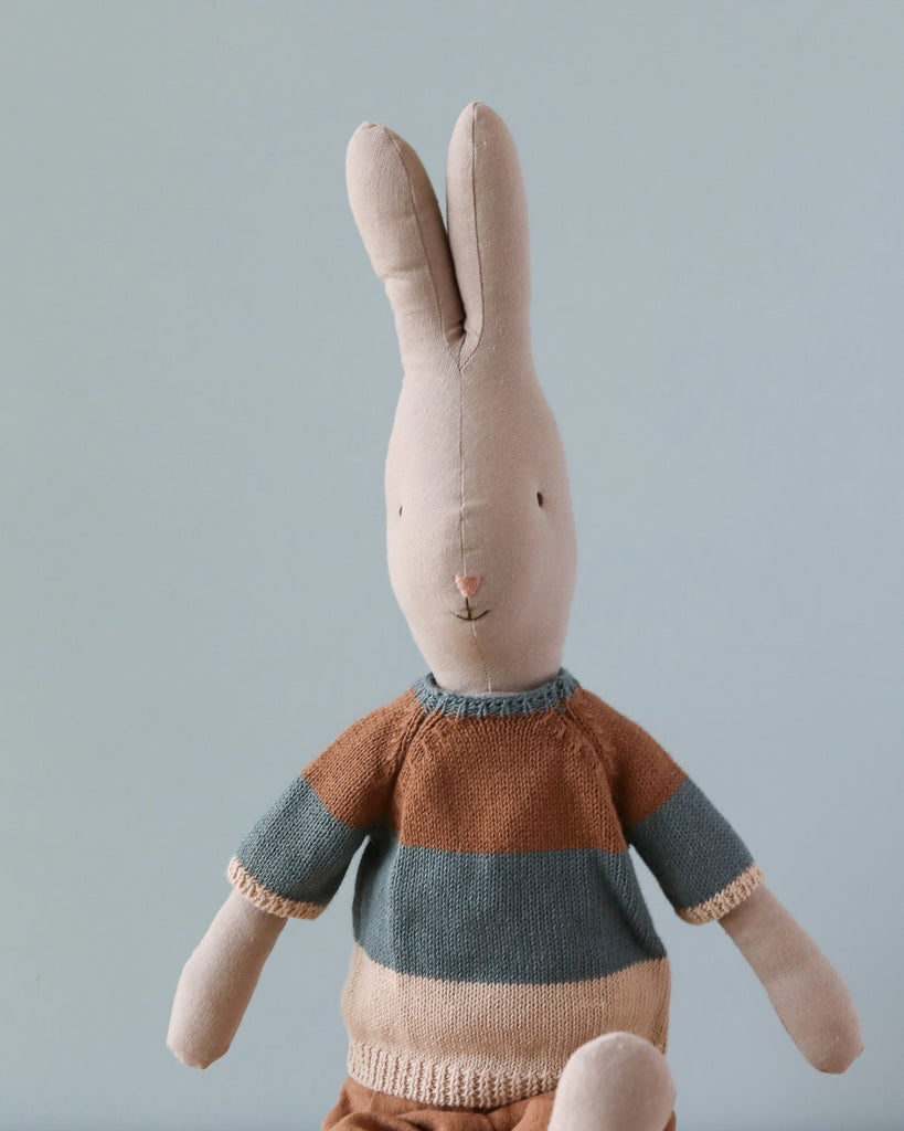 The Maileg Rabbit Size 4, Classic - Knitted Shirt & Shorts is seated against a plain light blue background. This plush toy rabbit, with long ears, wears a striped sweater in shades of blue and brown. Made from natural linen and cotton fabric, the minimalist bunny features small stitched eyes and a tiny pink nose—ideal for any bunny and rabbit collection.