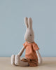 A Maileg Rabbit Size 1 - Classic T-shirt & Shorts sits upright on a wooden surface against a light blue background. The rabbit, made from recycled polyester, is wearing an orange and white striped shirt and brown shorts. Its ears point up, and it has a neutral expression on its face.