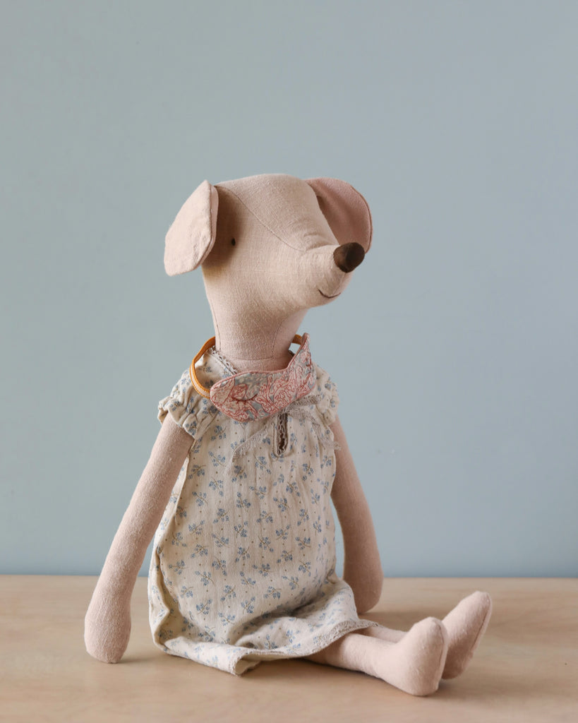 A Maileg Maxi Mouse in Nightgown sits on a wooden surface against a plain light blue background. The mouse is dressed in a light-colored floral nightgown and a decorative fabric collar. Its soft cotton/rime body and floppy ears give it a charming and whimsical appearance.