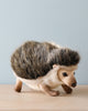 A realistic Hedgehog Stuffed Animal on a wooden surface against a light blue background. The hedgehog is detailed, capturing its spines, small eyes, and tiny paws in a natural color.