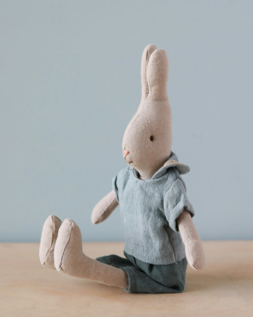 A beige fabric Maileg Rabbit, Size 2 - Classic Shirt & Shorts made from soft natural linen sits upright against a light blue background. The rabbit, perfect for a Maileg dollhouse, wears a light blue shirt and matching shorts, with its legs extended in front. The toy has stitched facial features, exuding a cute and minimalist charm.