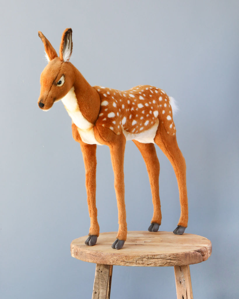 A Large Deer Stuffed Animal - Female with brown fur, white spots, dark hooves, and large ears stands upright on a small wooden stool against a plain gray background. Its realistic features make it stand out among other hand sewn animals.