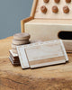 A wooden toy credit card and coins on a table, with a Wooden Cash Register in the background, simulating a finance or banking scenario for educational play.