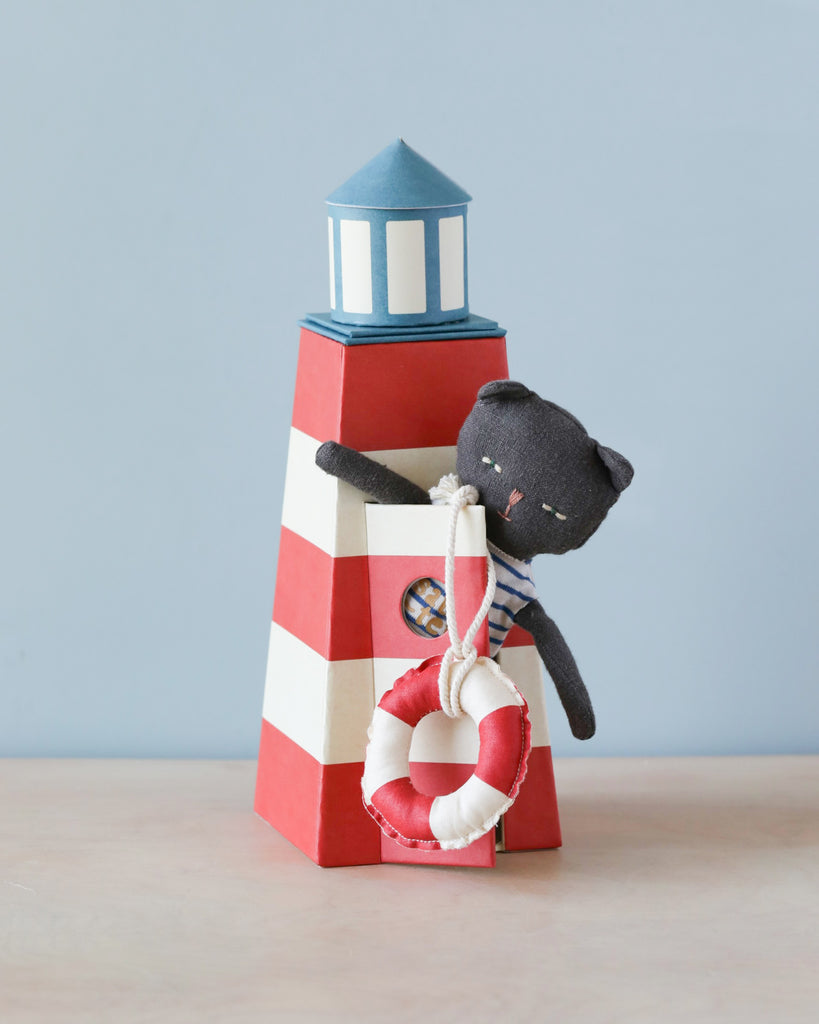 A Maileg Cat in Tower with red and white stripes, featuring a small window and a blue top, stands on a wooden surface. A fabric cat toy dressed as a sailor peeks from the window, while a toy lifebuoy, reminiscent of those used by lifeguards for swimmers in need, is attached to the tower.