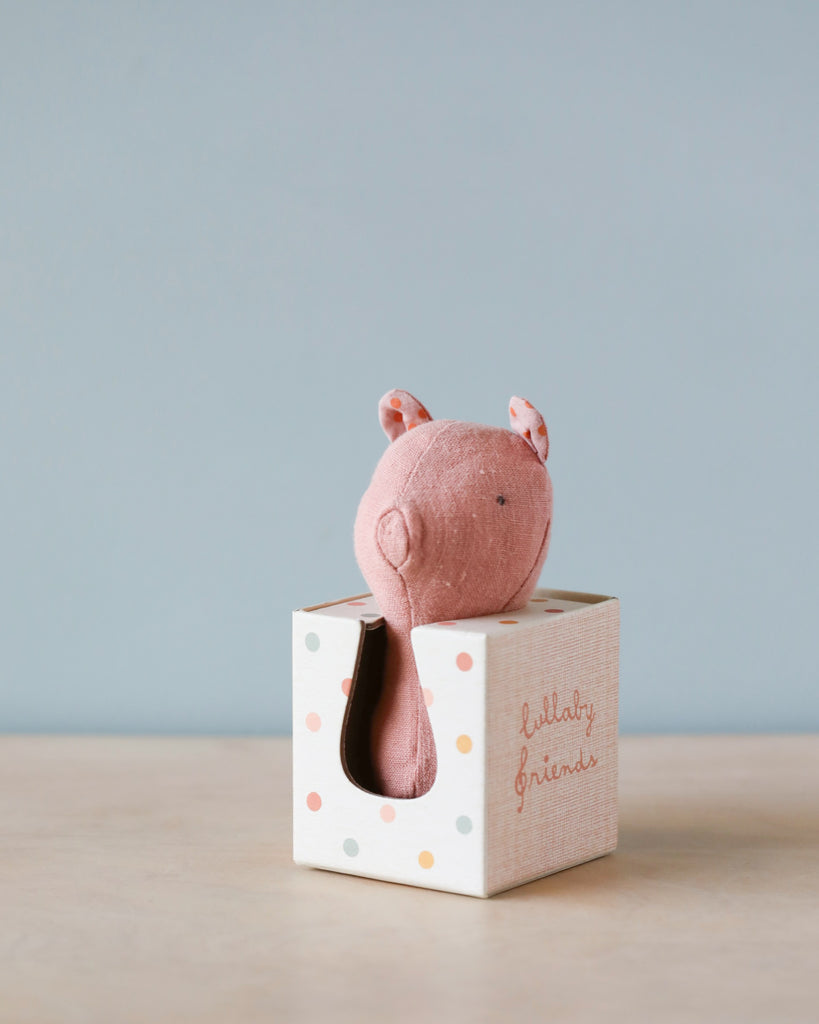 A cute plush toy resembling a pink bunny peeks out from a pastel-colored, polka dot gift box on a wooden surface. The box has the words "Maileg Lullaby Friend Rattles, Pig" written on it and is made of soft fabrics. The background is a light blue wall.