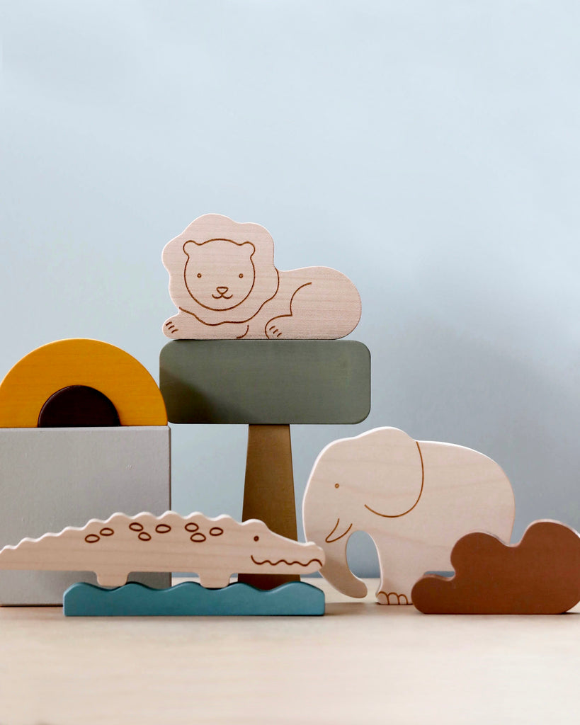 Wooden Safari Set, featuring hand-painted maple wood animals like a lion, elephant, and crocodile, are arranged on and around colorful blocks. The lion sits atop a green block, the elephant stands beside the blocks, while the crocodile rests near the base. A cloud-shaped cutout is also seen. These toys aid in fine motor skills development.