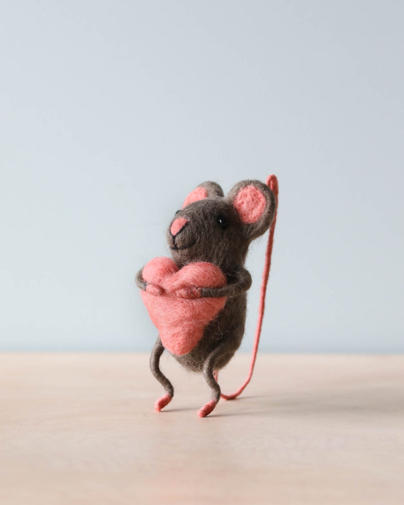 A small hand-felted grey mouse holding a pink heart in its arms. 